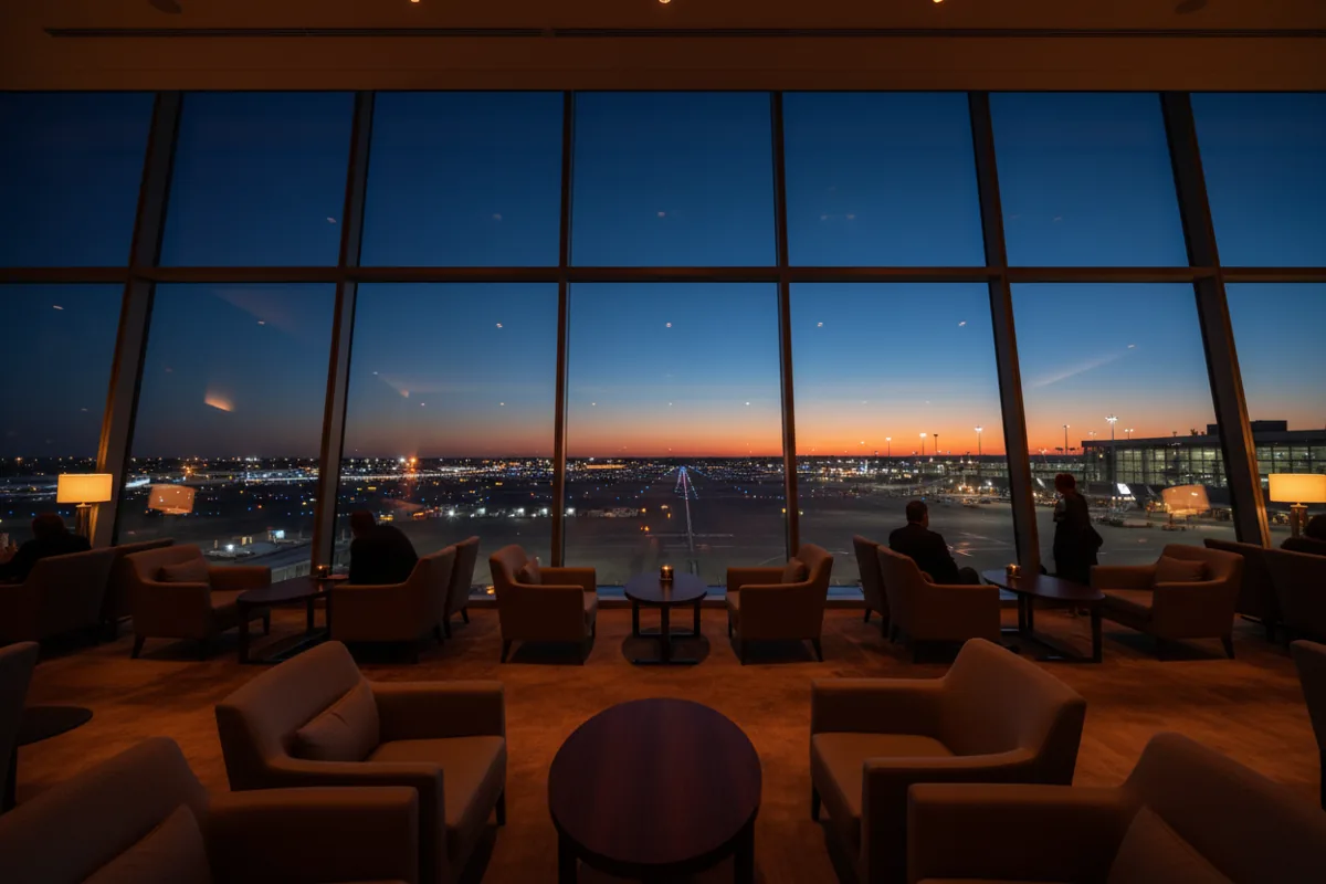The interior of a modern, empty airport lounge with a view of the tarmac at dusk.