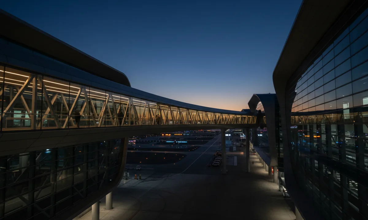 An illuminated pedestrian sky bridge connecting parts of a modern airport terminal at blue hour.