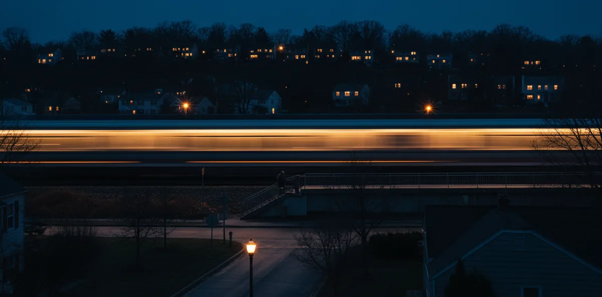 A commuter train shown as a streak of light as it travels through a suburban town at dusk.
