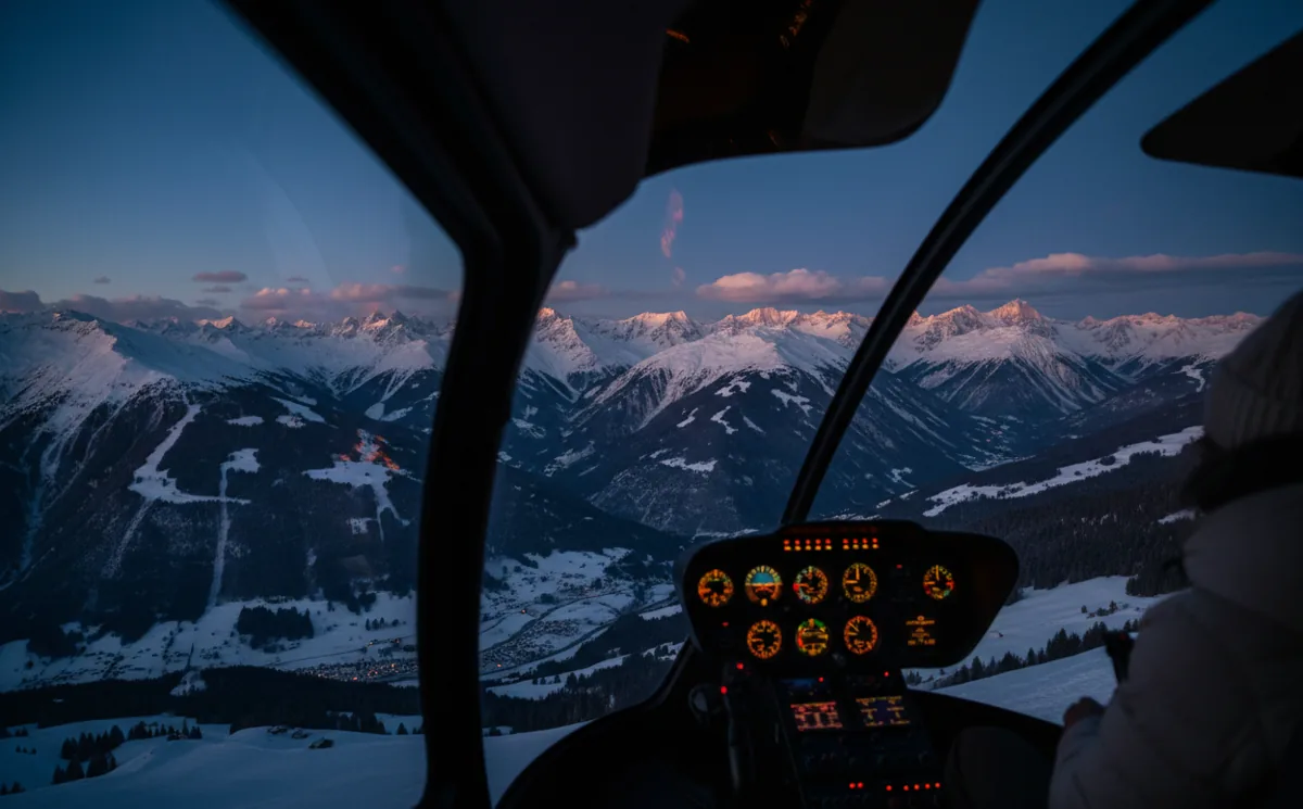View from a helicopter window of snow-covered Alpine peaks at sunset.