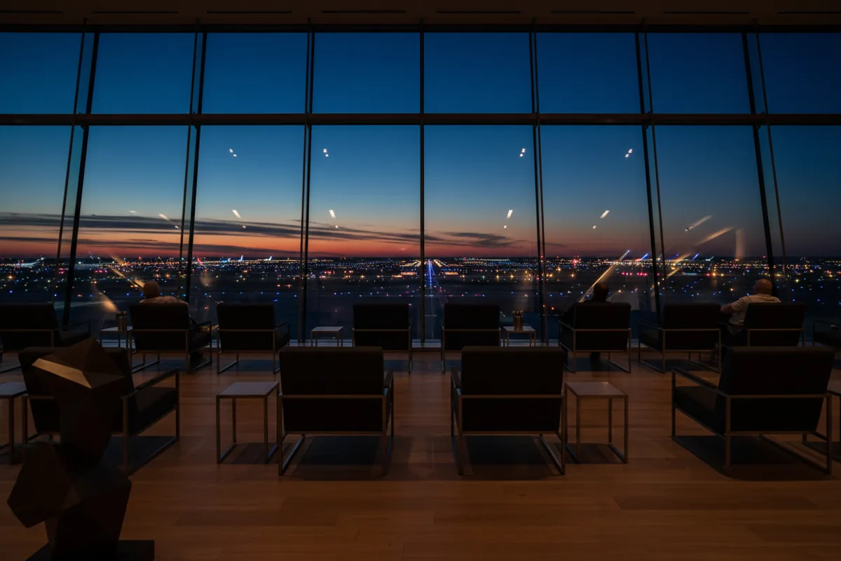 The interior of a modern airport lounge with large windows looking out onto the airfield at dusk.