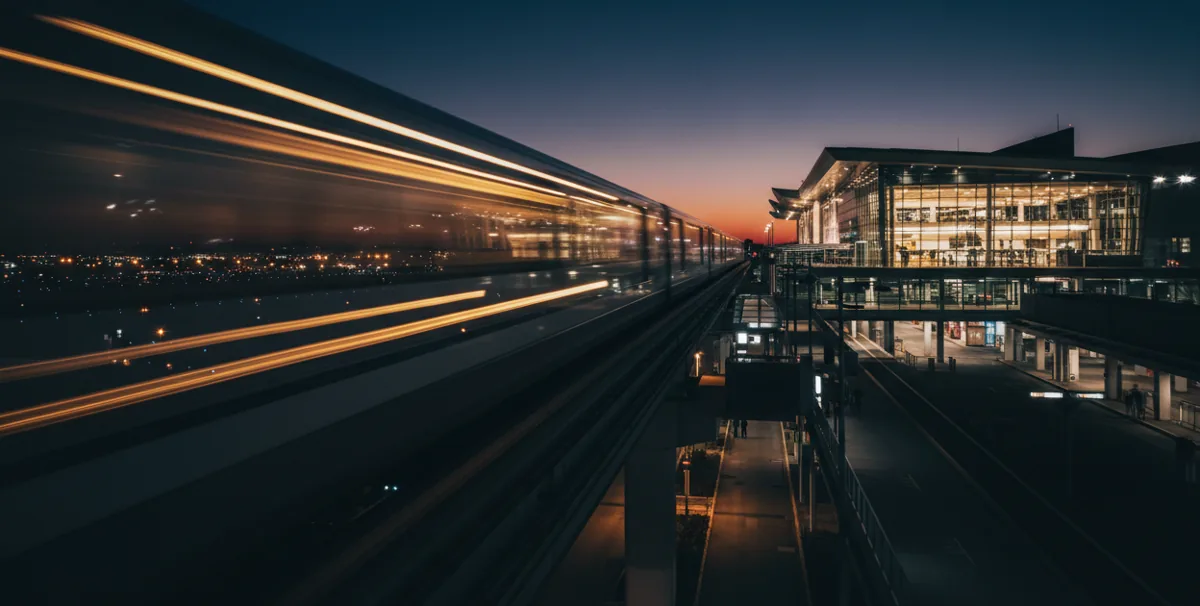 An elevated airport train in motion at night, approaching a brightly lit terminal building.