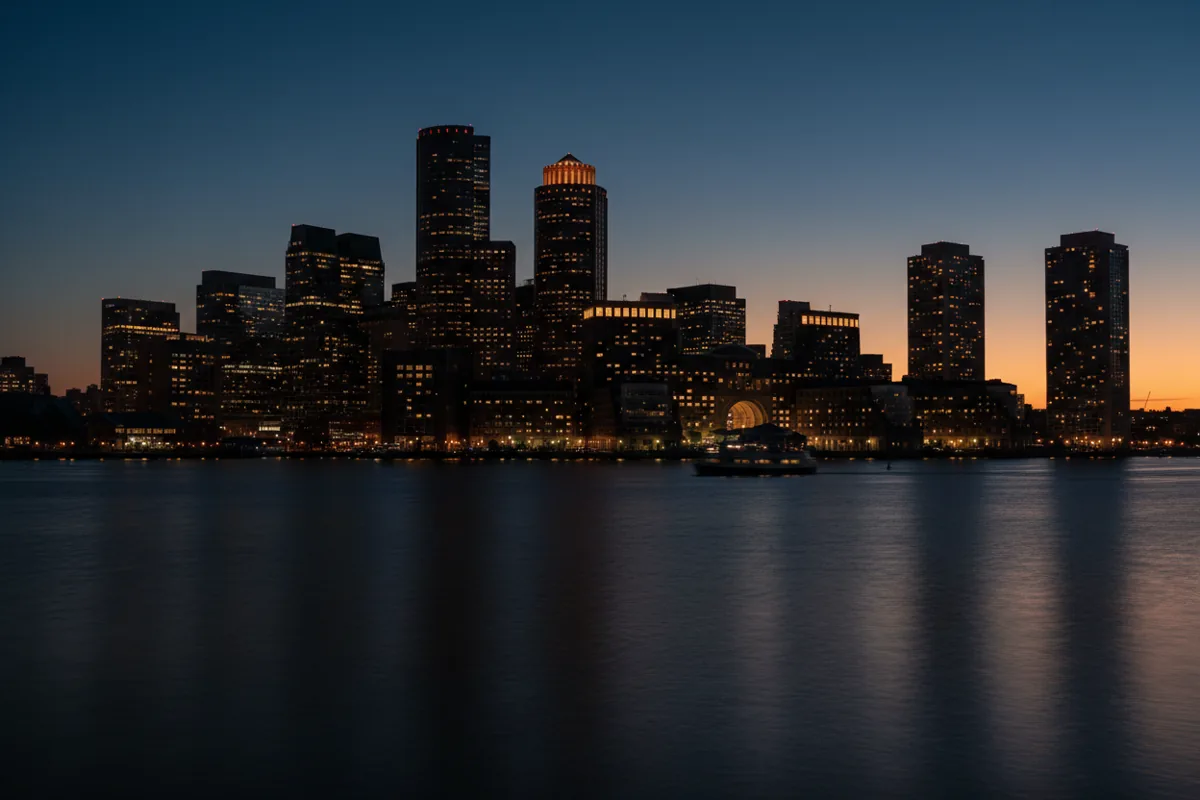 The Boston city skyline seen across the water at twilight, with lights glowing in the buildings.