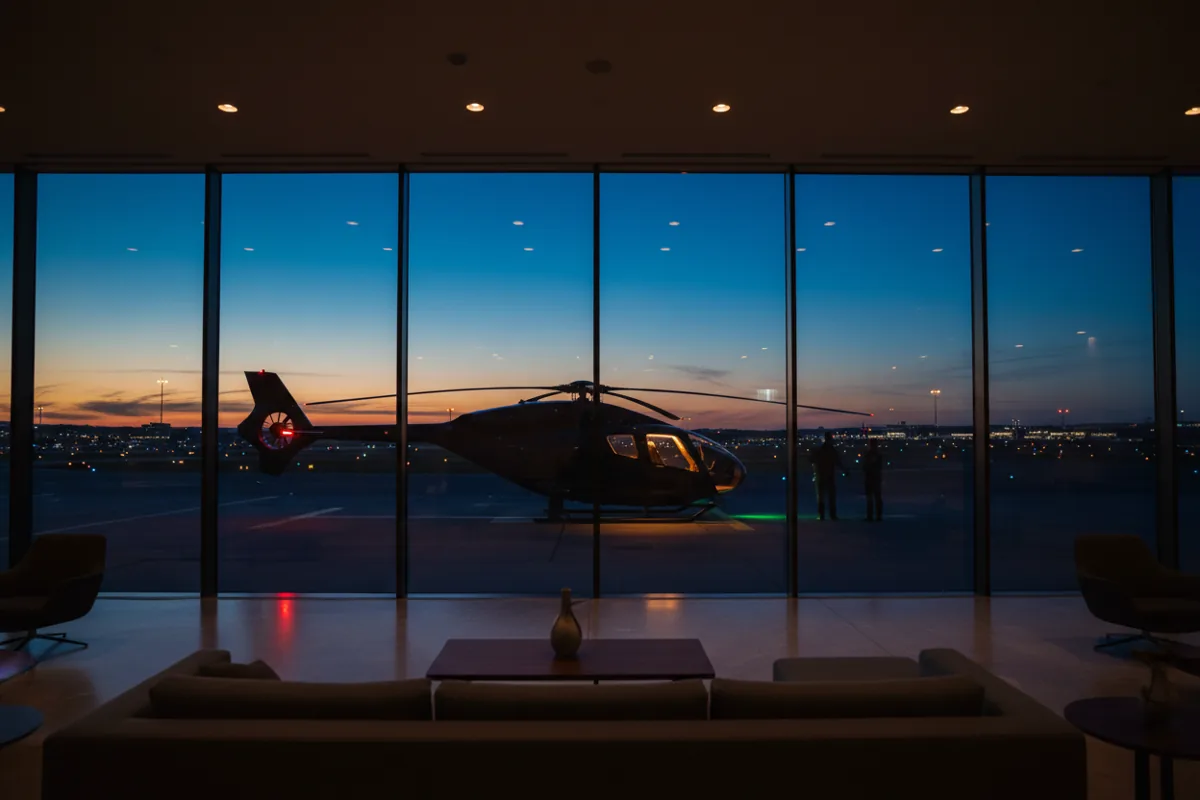 A helicopter sits on a helipad at dusk, viewed from inside a modern airport lounge.