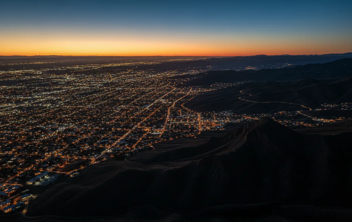 Aerial view at dusk of city lights spreading towards the dark foothills of a mountain range.