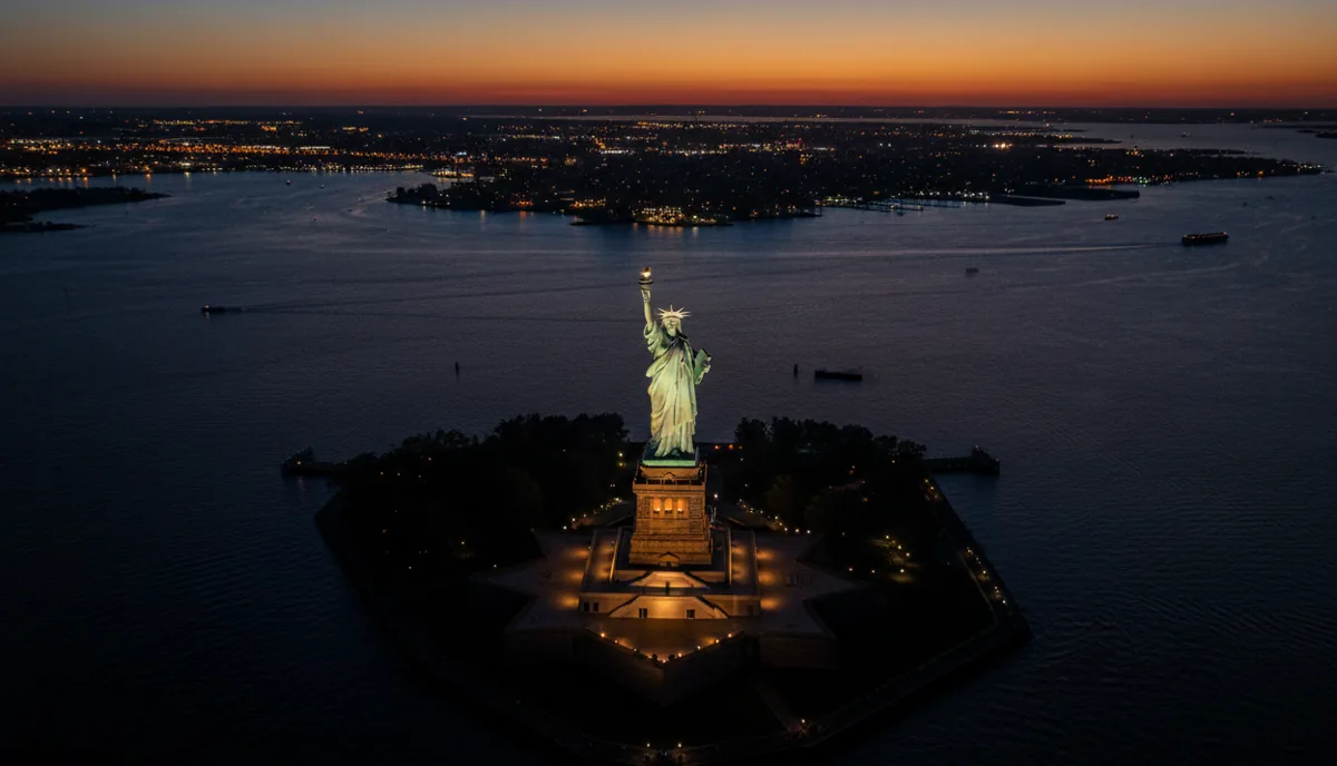 Aerial view of the illuminated Statue of Liberty at night, with city lights in the distance.