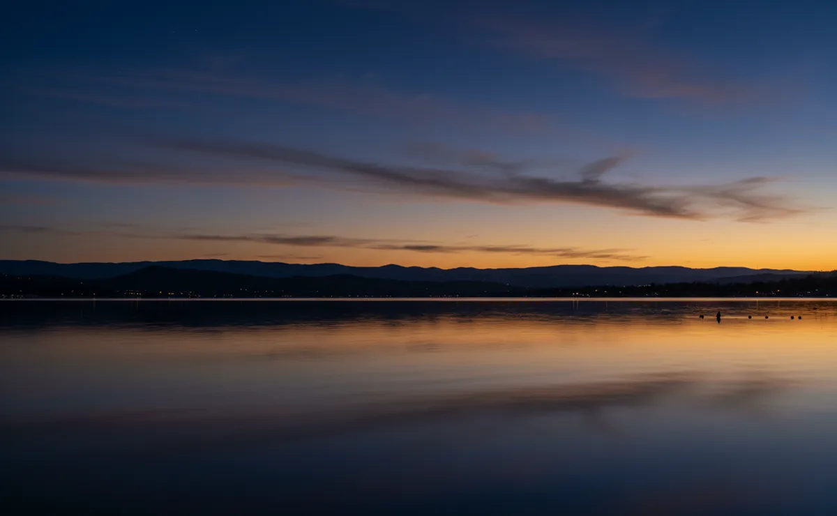The calm water of Folsom Lake at dusk, reflecting a purple and orange sunset sky over distant foothills.