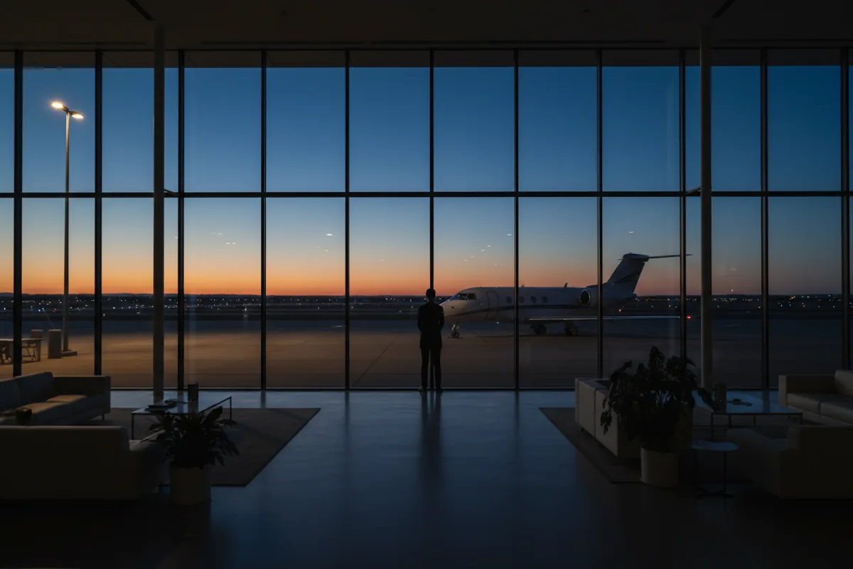 A person in silhouette looks out from a modern airport lounge onto a private jet on the tarmac at twilight.