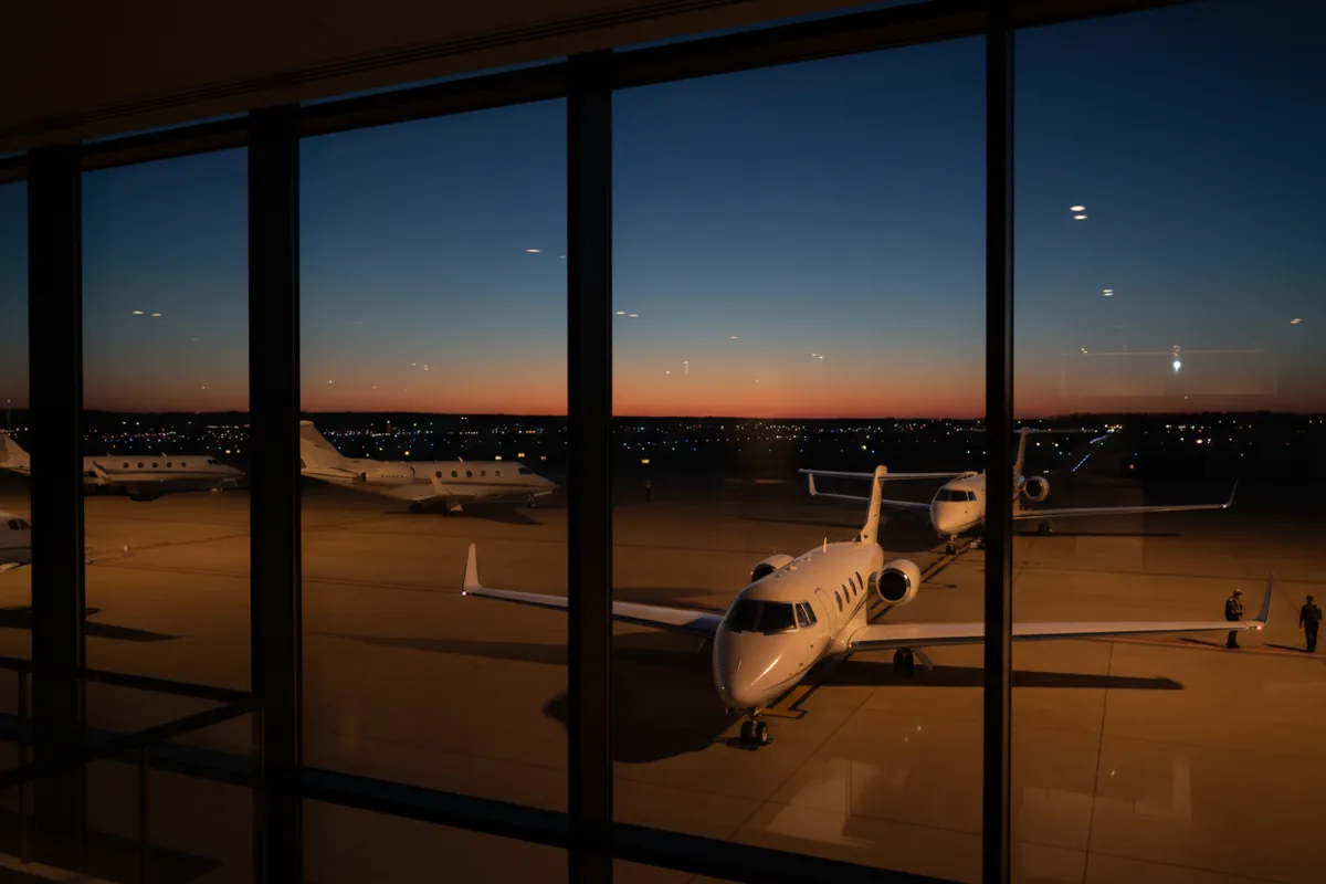 Unmarked private jets parked on an airport tarmac at dusk, viewed from inside the terminal.