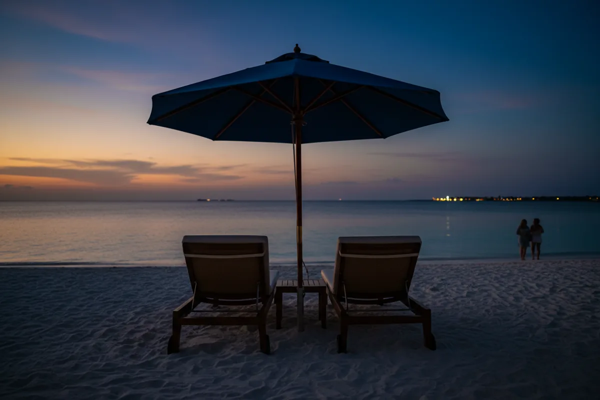 Two empty lounge chairs and an umbrella on a white sand beach facing a calm turquoise ocean.