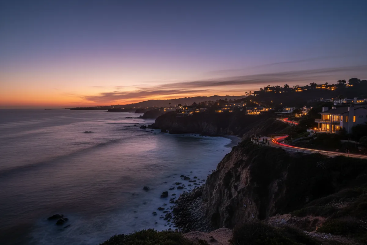 A dramatic coastline view at dusk with glowing lights from homes on the cliffs above the ocean.