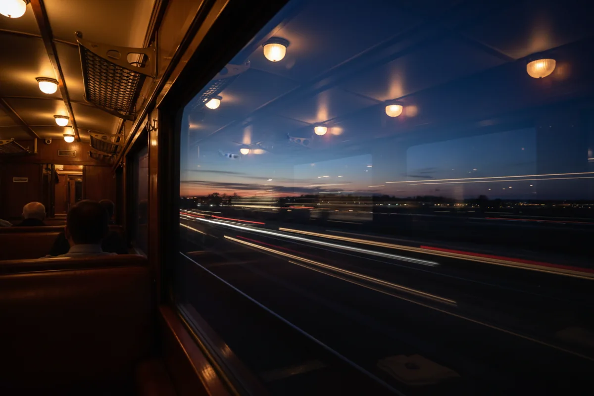 View from a train window at night, showing motion-blurred lights outside and a reflection of the warm interior.