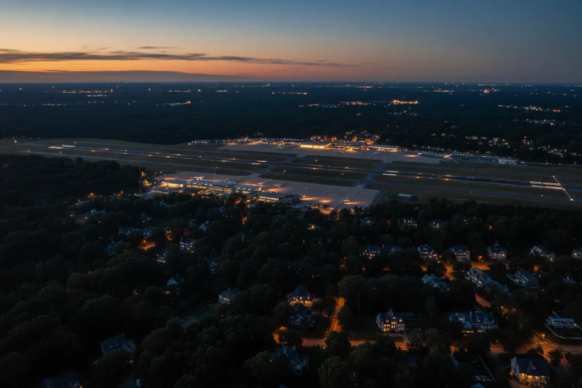 Aerial view of an airport's glowing runways at night, surrounded by dark woods and suburban lights.