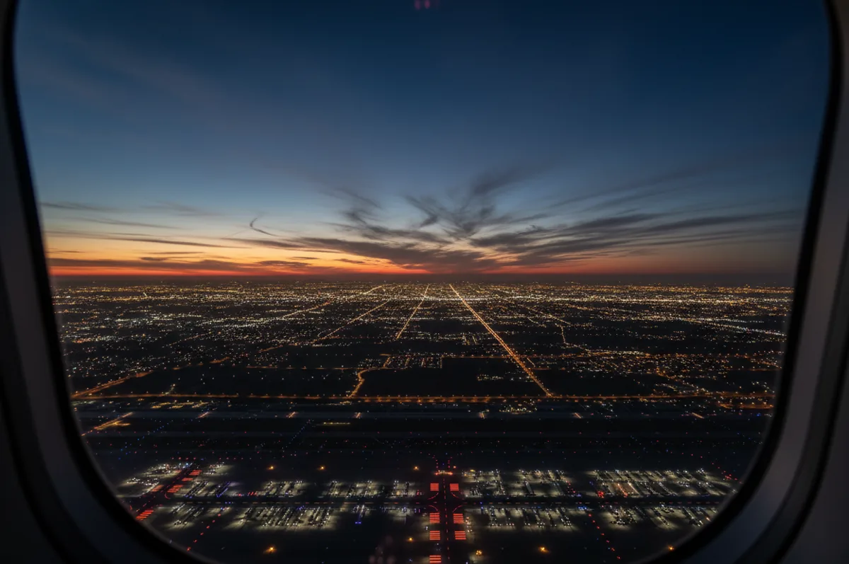 View from a departing airplane at night, looking down on airport lights and a sprawling city grid on the horizon.
