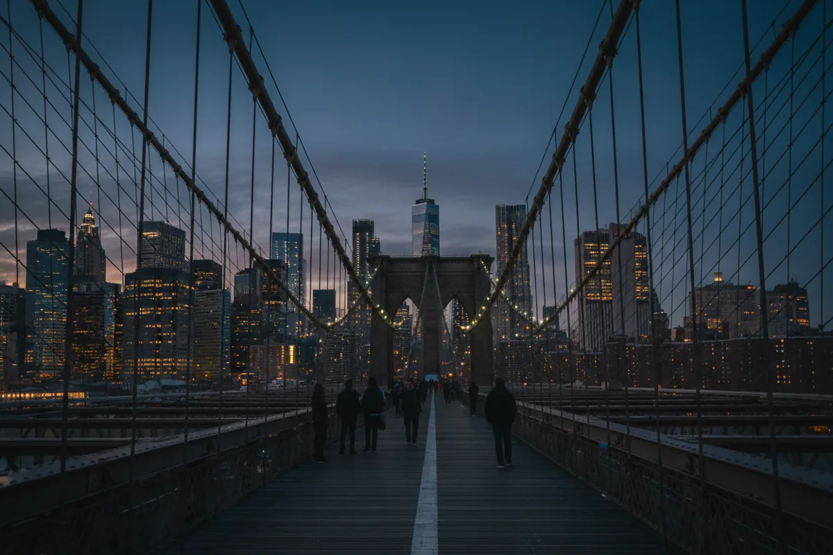 The Brooklyn Bridge walkway at dusk, with its cables framing the illuminated Lower Manhattan skyline.