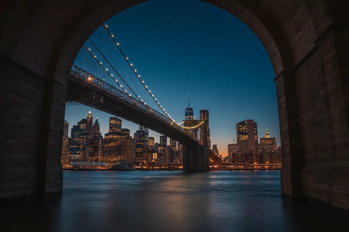 The Manhattan skyline viewed through the arches of the Brooklyn Bridge at twilight.